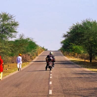 Motorcyclist riding down a rural road in Rajasthan, India, with two people in traditional Indian clothing walking on the side.