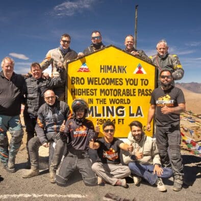Motorcyclists celebrating at Umling La, the highest motorable pass in the Himalayas, with the official sign and prayer flags in the background.