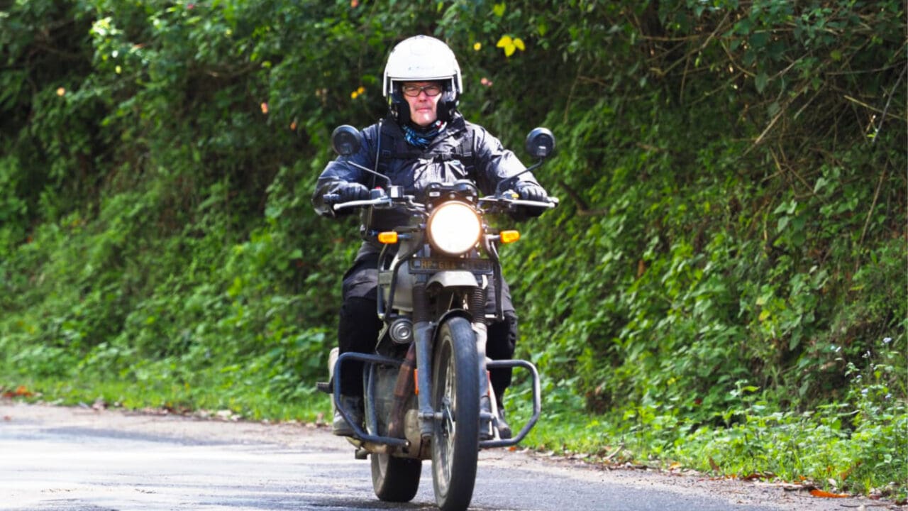 A motorcyclist with protective gear riding on a motorcycle along a green forested road in South India.