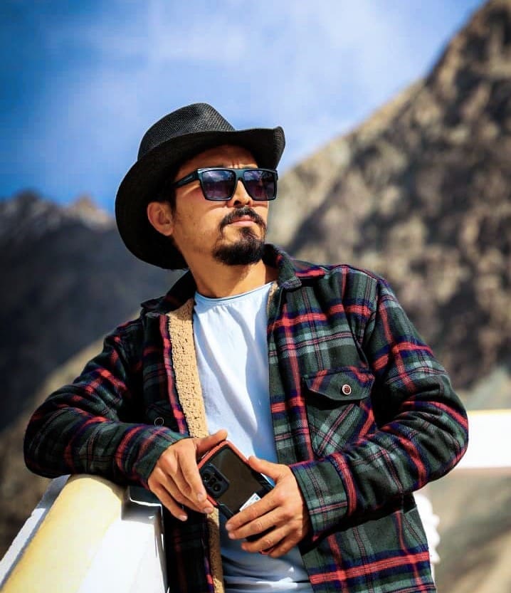Adventure tour guide Nubu Sunny wearing a cowboy hat and sunglasses, holding a smartphone, with a mountain range in the background.