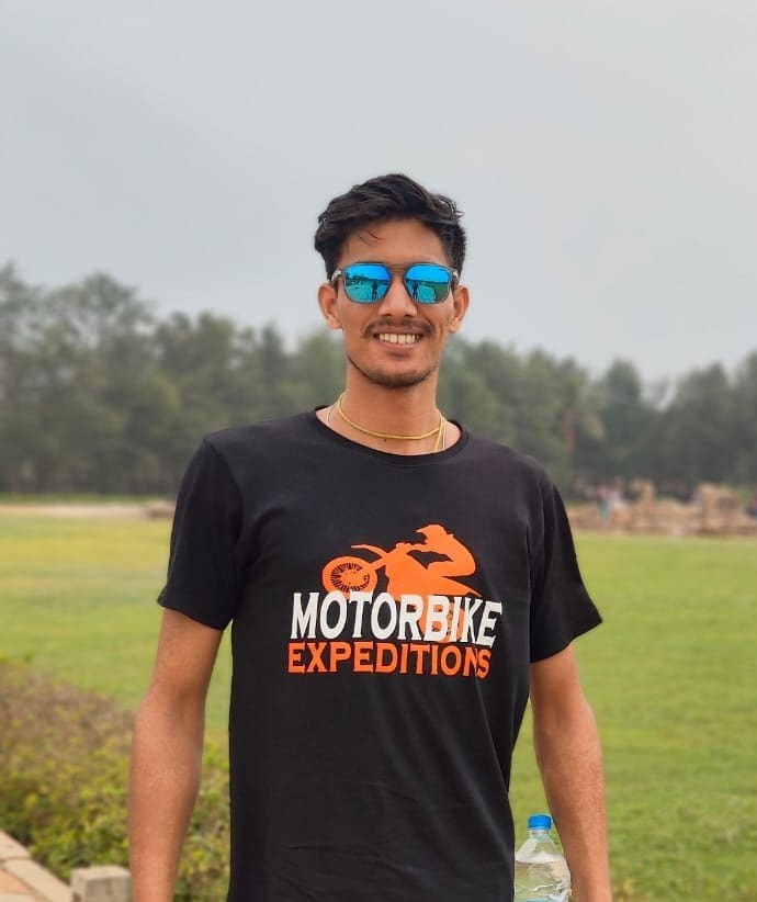 Enthusiastic motorbike tour guide Manish in a black expedition-themed t-shirt, posing with blue sunglasses in a natural outdoor setting.