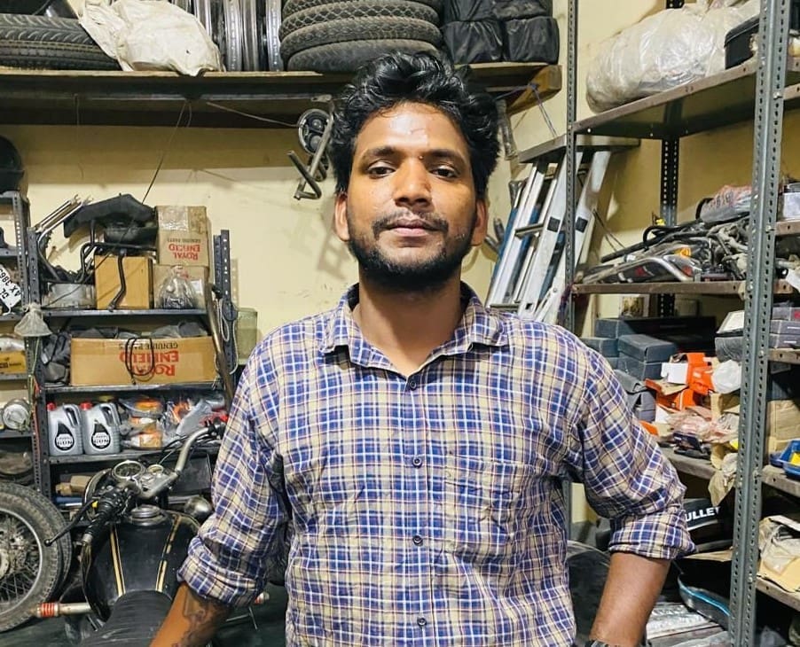 Tour mechanic named Sattu standing in a motorbike repair shop with shelves of bike parts and tools in the background.