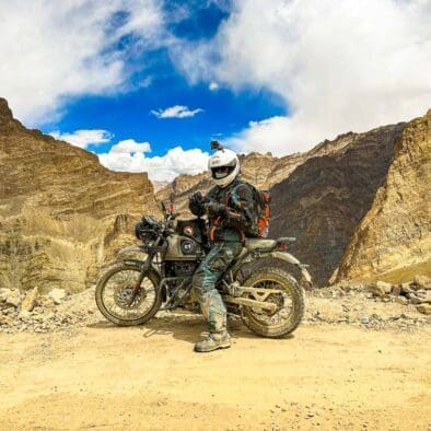 Motorcyclist in full gear standing with a touring motorcycle on a Himalayan dirt road with mountainous terrain in the background.