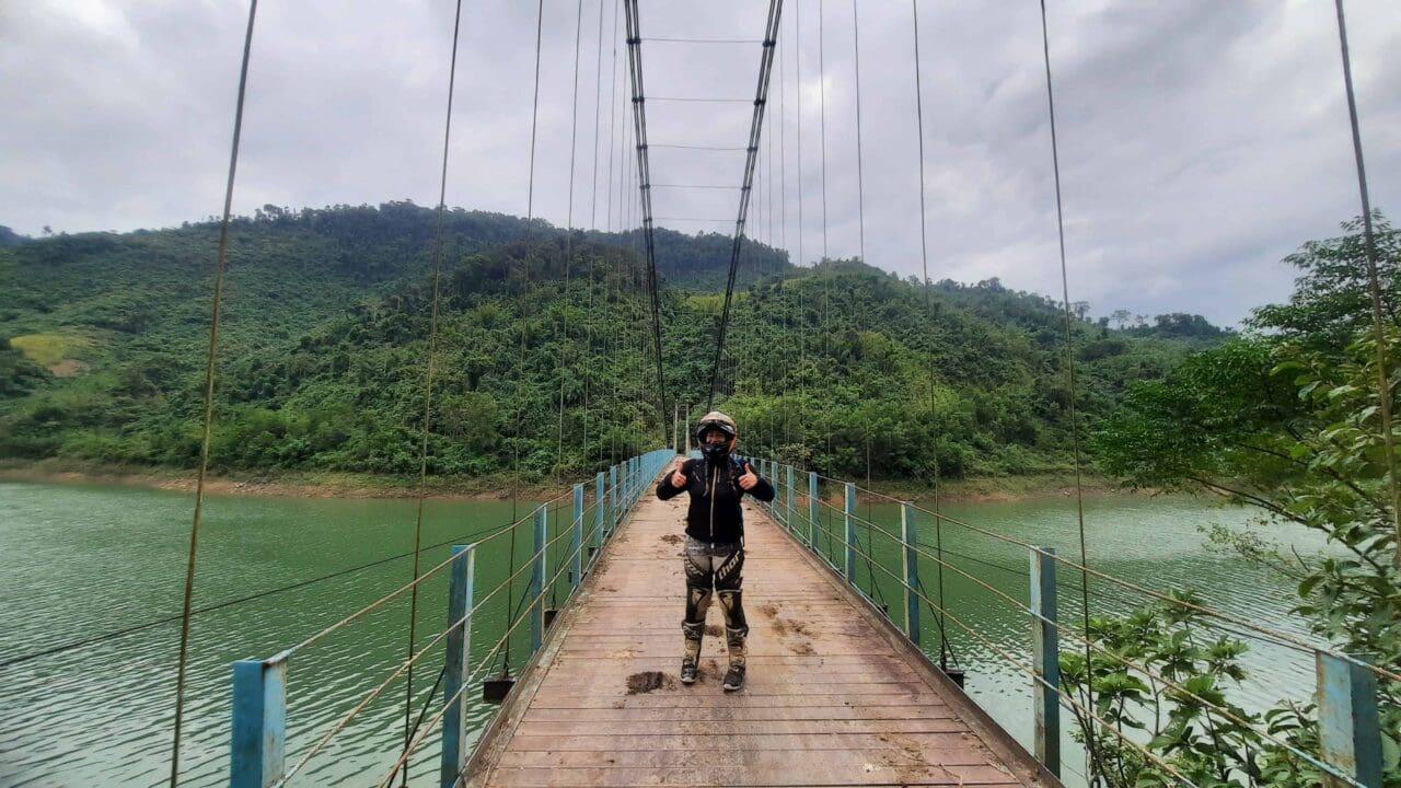 Traveler in full gear giving thumbs up while crossing a suspension bridge over a river in a lush, green mountainous area of Central Vietnam.