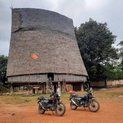 Two motorcycles parked in front of a traditional Vietnamese Rong house with a thatched roof, surrounded by trees.