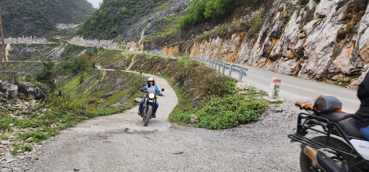 Female motorcyclist riding on a narrow mountain road in North Vietnam, with steep cliffs and lush greenery in the background.