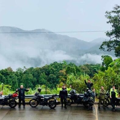 Group of motorbike riders posing on a wet road with misty mountains in the background.