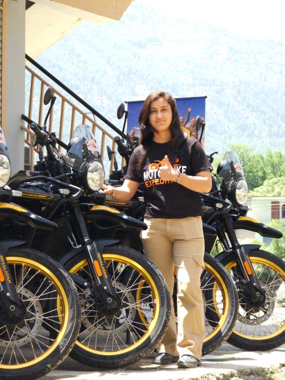 Mohini, office manager at Motorbike Expeditions Ltd, wearing a black t-shirt and beige pants, standing beside motorbike.