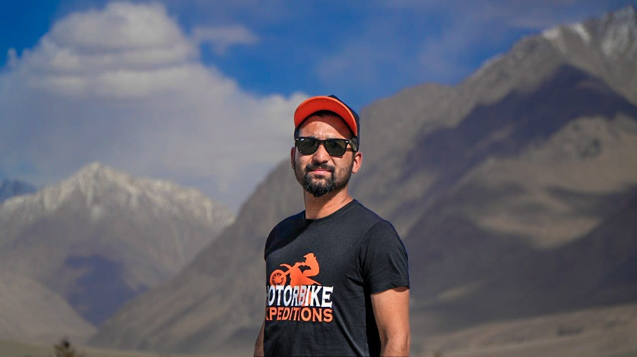 Surya Thakur, co-founder of Motorbike Expeditions Ltd, wearing a black t-shirt and orange cap, with mountains in the background.